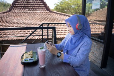 Genç Asyalı tesettürlü bir kadın, bir kafede dinlenirken cep telefonundan bir müzik çalarıyla müzik dinlemekten zevk alıyor. Teknoloji konsepti..