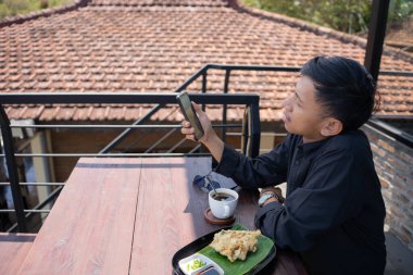 Genç Asyalı bir adam cep telefonuyla oynarken kafenin balkonunda dinleniyor ve bir fincan sade kahve ve bir tabak kızarmış tempeh ısmarlıyor..