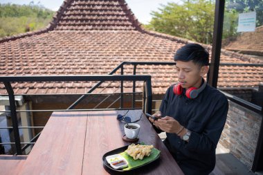 Genç Asyalı bir adam cep telefonuyla oynarken kafenin balkonunda dinleniyor ve bir fincan sade kahve ve bir tabak kızarmış tempeh ısmarlıyor..