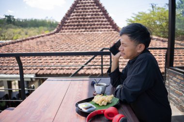 a handsome young Asian man was making a call on his cell phone while hanging out at a cafe. clipart