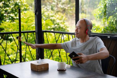 a young bald Asian man is listening to music with his earphones and seems to be enjoying it while hanging out and relaxing at a cafe. clipart