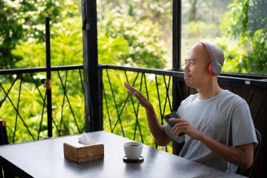 a young bald Asian man is listening to music with his earphones and seems to be enjoying it while hanging out and relaxing at a cafe. clipart