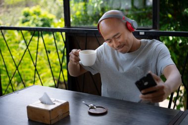 Genç ve kel bir Asyalı adam kulaklıklarıyla müzik dinliyor ve kahve içerken bir kafede dinleniyor gibi görünüyor..