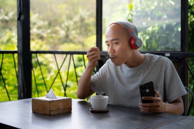 Genç ve kel bir Asyalı adam kulaklıklarıyla müzik dinliyor ve bir kafede takılıp dinlenirken bundan zevk alıyor gibi görünüyor..