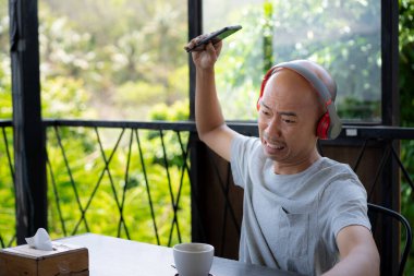 a bald Asian youth is playing a game on his smartphone and looks angry and frustrated while hanging out and relaxing at a cafe. clipart