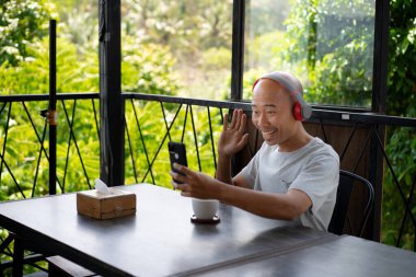 A bald Asian boy looks happy as he receives a video call on his smartphone while hanging out at a cafe. clipart