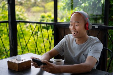 Kel bir Asyalı genç akıllı telefonuyla oyun oynuyor ve bir kafede takılıp dinlenirken mutlu görünüyor..