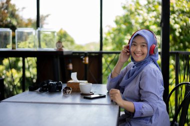 young Asian woman in hijab enjoying music using a headsets with a music player from her mobile phone while relaxing in a cafe, technology concept. clipart