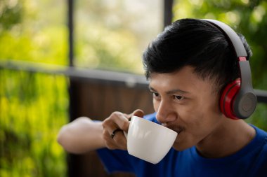 Bir kafede kahve içen yakışıklı genç bir Asyalı mutlu görünüyor ve akıllı telefonuyla kulaklık takıp şarkı dinlemekten hoşlanıyor..