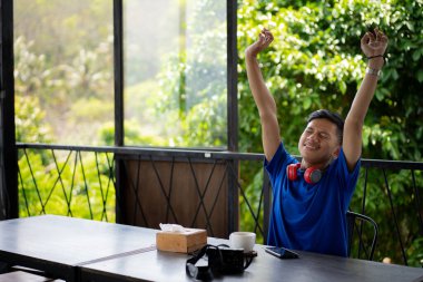 Mavi tişörtlü, sıkılmış genç Endonezyalı bir adam bir kafede oyun oynarken kollarını kaldırıyor..