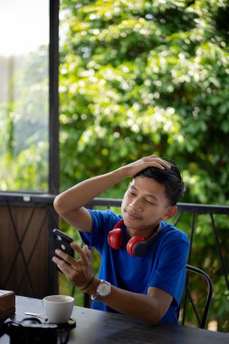 A young Indonesian man in a blue shirt looks sad when he loses when playing a cellphone game at a cafe. clipart