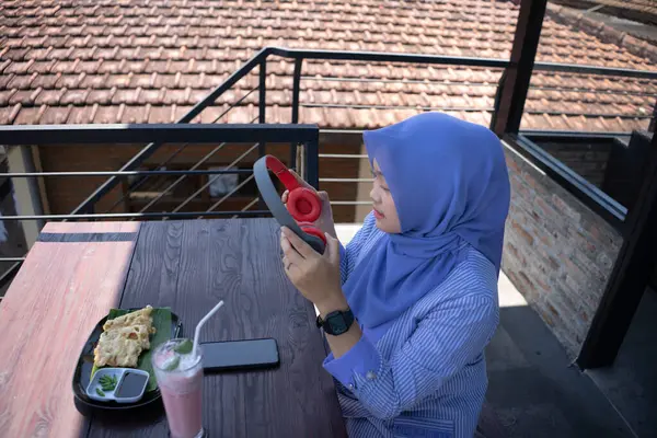 Genç Asyalı tesettürlü bir kadın, bir kafede dinlenirken cep telefonundan bir müzik çalarıyla müzik dinlemekten zevk alıyor. Teknoloji konsepti..