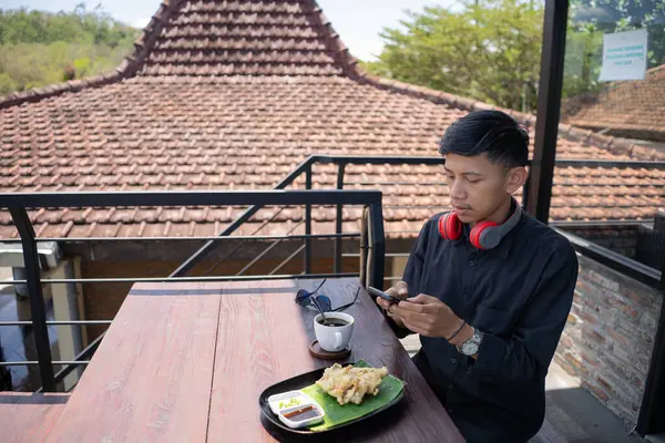 Genç Asyalı bir adam cep telefonuyla oynarken kafenin balkonunda dinleniyor ve bir fincan sade kahve ve bir tabak kızarmış tempeh ısmarlıyor..