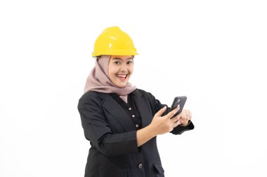 Young Indonesian businesswoman wearing hijab and safety hat standing confidently with cellphone looking at camera, concept of women boss and industry, isolated on white background. clipart