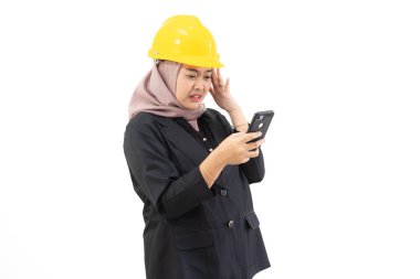 Young Indonesian businesswoman wearing hijab and safety hat standing confidently with cellphone looking at camera, concept of women boss and industry, isolated on white background. clipart