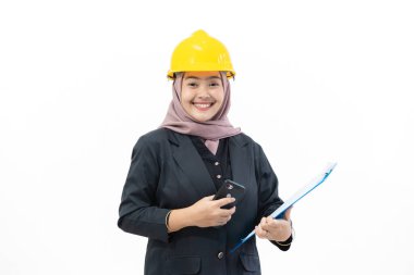 Genç Endonezyalı kadın patron, tesettür ve güvenlik şapkası takıyor. Güvenle kameraya bakıyor, pano ve cep telefonu kullanıyor. Kadın patron ve endüstri kavramı beyaz arka planda izole edilmiş..