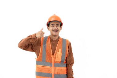Endonezyalı genç inşaat işçisi tam güvenlik teçhizatıyla kameraya, endüstriyel ve inşaat konseptine, izole edilmiş beyaz arka plana başparmağıyla güvenle duruyor..