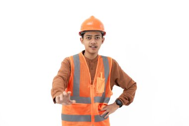 Endonezyalı genç inşaat işçisi, güvenlik teçhizatıyla kameraya, inşaata ve sanayi konseptine işaret ederek beyaz arka planda izole bir şekilde el hareketi yapıyor..