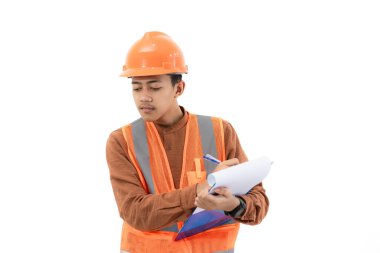 Tam güvenlik donanımlı Endonezyalı genç inşaat işçisi, beyaz arka planda izole edilmiş bir pano, inşaat ve sanayi iş konsepti hakkında bir rapor doldurmaya odaklanırken görülüyor..