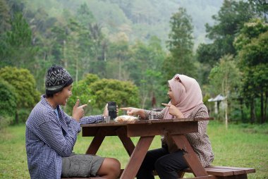 Endonezyalı genç kadınlar tesettür takıyor ve Endonezyalı genç erkekler piknikte piknik yaparken cep telefonlarına bakıyorlar..