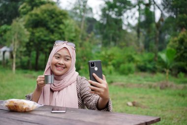 Genç Endonezyalı tesettürlü kadın sade kahve içerken piknik yaparken sakinleştirici doğal atmosferin ve seyahat konseptinin tadını çıkarıyor..