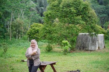 Genç Endonezyalı kadın tesettüre binmiş, rahatlatıcı doğal atmosferin tadını çıkarırken bir fincan sade kahvenin tadını çıkarıyor, şehrin koşuşturma ve telaşından uzakta, seyahat konseptinden uzakta..