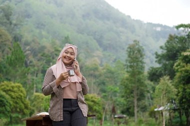 Genç Endonezyalı kadın tesettüre binmiş, rahatlatıcı doğal atmosferin tadını çıkarırken bir fincan sade kahvenin tadını çıkarıyor, şehrin koşuşturma ve telaşından uzakta, seyahat konseptinden uzakta..