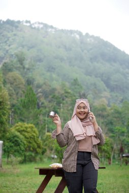Genç Endonezyalı kadın tesettüre binmiş, rahatlatıcı doğal atmosferin tadını çıkarırken bir fincan sade kahvenin tadını çıkarıyor, şehrin koşuşturma ve telaşından uzakta, seyahat konseptinden uzakta..