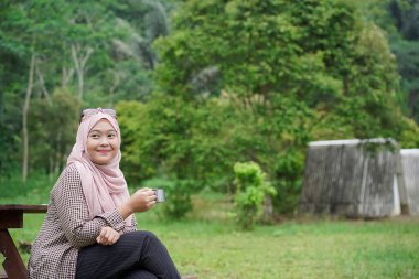 Genç Endonezyalı kadın tesettüre binmiş, rahatlatıcı doğal atmosferin tadını çıkarırken bir fincan sade kahvenin tadını çıkarıyor, şehrin koşuşturma ve telaşından uzakta, seyahat konseptinden uzakta..