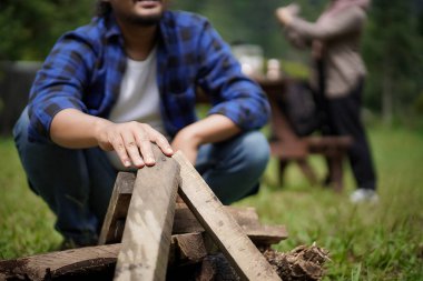 Endonezyalı adam kamp ateşi yakmak için odun hazırlarken Endonezyalı genç kadın tesettür ve seyahat konseptiyle doğada kamp yapıyor..