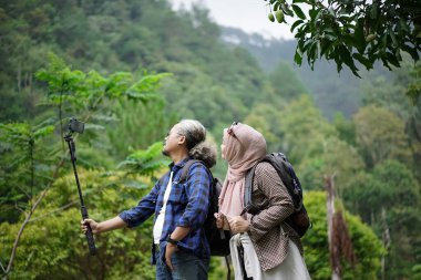 Kıvırcık saçlı Endonezyalı bir adam ve tesettürlü genç bir kadın ormanda yürürken video çekip doğal manzaranın ve seyahat konseptinin tadını çıkarıyorlar..