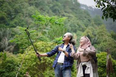 Kıvırcık saçlı Endonezyalı bir adam ve tesettürlü genç bir kadın ormanda yürürken video çekip doğal manzaranın ve seyahat konseptinin tadını çıkarıyorlar..