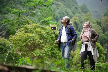Kıvırcık saçlı Endonezyalı bir adam ve tesettürlü genç bir kadın ormanda yürürken video çekip doğal manzaranın ve seyahat konseptinin tadını çıkarıyorlar..