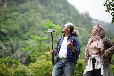 Kıvırcık saçlı Endonezyalı bir adam ve tesettürlü genç bir kadın ormanda yürürken video çekip doğal manzaranın ve seyahat konseptinin tadını çıkarıyorlar..
