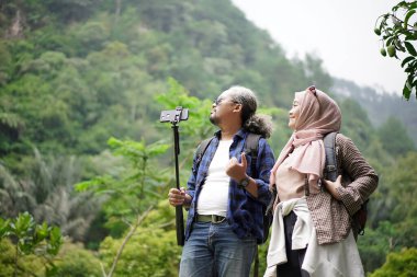 Kıvırcık saçlı Endonezyalı bir adam ve tesettürlü genç bir kadın ormanda yürürken video çekip doğal manzaranın ve seyahat konseptinin tadını çıkarıyorlar..