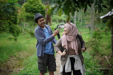 Endonezyalı genç bir adam, Endonezyalı genç bir kadının doğada yürürken ve seyahat konseptinde video kayıt cihazları hazırlamasına yardım ediyor..