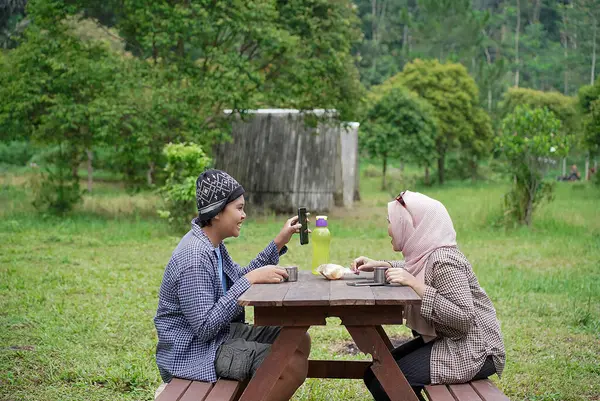 Endonezyalı genç kadınlar tesettür takıyor ve Endonezyalı genç erkekler piknikte piknik yaparken cep telefonlarına bakıyorlar..