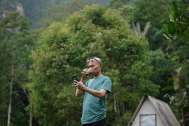 Kel kafalı genç Endonezyalı bir adam orman kulübesinde kamp yaparken bir fincan sade kahvenin tadını çıkarıyor..