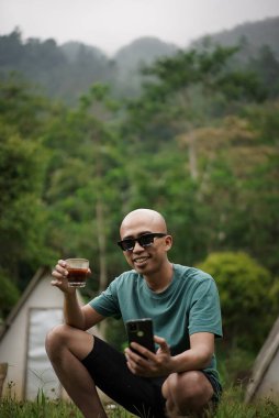 Kel kafalı genç Endonezyalı gözlüklü adam orman kulübesinde cep telefonu taşırken bir fincan sade kahvenin tadını çıkarıyor. Seyahat konsepti..