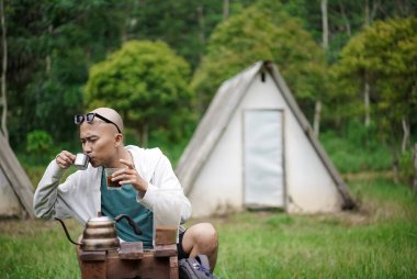 Kel kafalı Endonezyalı adam orman kulübesinde kamp yaparken bir fincan ev yapımı sade kahveden hoşlanıyor. Seyahat kavramı..