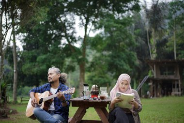Endonezyalı erkek ve genç Endonezyalı kadın piknik masasında oturup birlikte orman kulübesinde kamp yaparken gitar çalıp kitap okuyorlar. Seyahat kavramı..