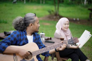 Endonezyalı erkek ve genç Endonezyalı kadın piknik masasında oturup birlikte orman kulübesinde kamp yaparken gitar çalıp kitap okuyorlar. Seyahat kavramı..