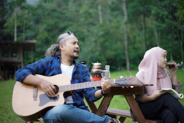 Endonezyalı erkek ve genç Endonezyalı tesettürlü kadın piknik masasında birlikte şarkı söyleyip gitar çalarken orman kulübesinde kamp yapıyorlar..