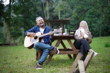 Endonezyalı erkek ve genç Endonezyalı tesettürlü kadın piknik masasında birlikte şarkı söyleyip gitar çalarken orman kulübesinde kamp yapıyorlar..