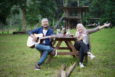 Endonezyalı erkek ve genç Endonezyalı tesettürlü kadın piknik masasında birlikte şarkı söyleyip gitar çalarken orman kulübesinde kamp yapıyorlar..