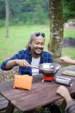 Kıvırcık saçlı Endonezyalı bir adam orman kulübesinde kamp yaparken dondurulmuş yemek pişiriyor. Kopya alanı ve reklamcılık, seyahat konsepti..