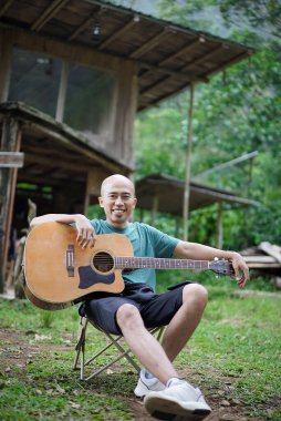 Endonezyalı kel adam orman kulübesinin önünde gitar çalıyor, seyahat konsepti..