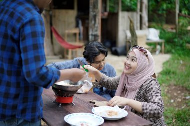 Endonezyalı genç bir adam ve kadın tesettürlü bir orman kulübesinin önünde dinleniyor gitar çalıyor ve donmuş yiyecek, seyahat konsepti kızartıyorlar..