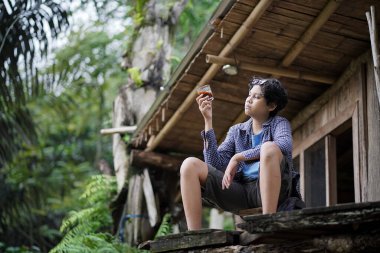 Genç Endonezyalı bir adam bir orman kulübesinde dinlenirken bir fincan kahvenin, seyahat konseptinin tadını çıkarıyor..