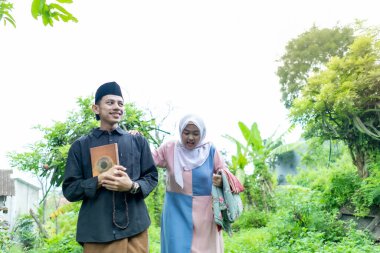 Bir çift genç Endonezyalı kadın ve erkek Müslüman kıyafetleri giyip Kuran 'a gidiyorlar. Kur 'an ve namaz araçlarını taşırlar..